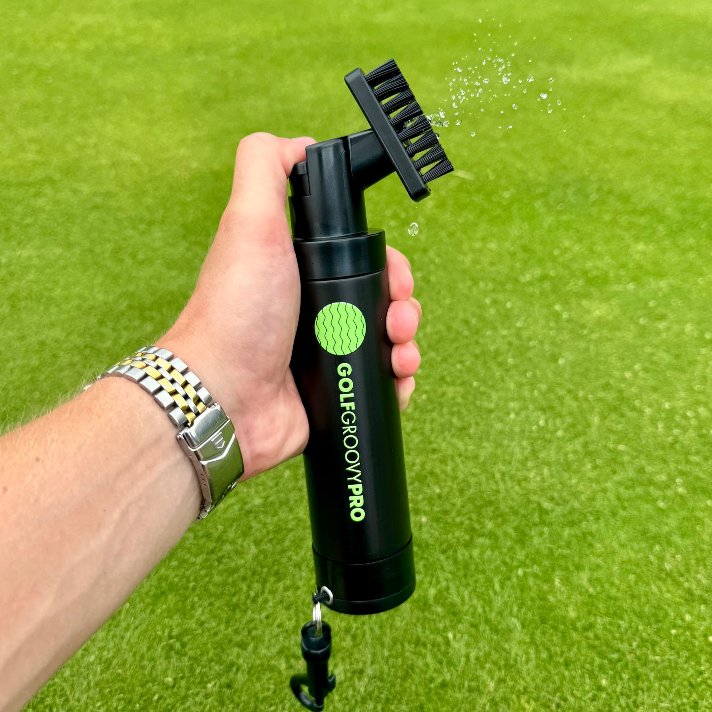 Hand holding a black and green cleaning tool labeled 'Golf Groovy Pro' on a grass background